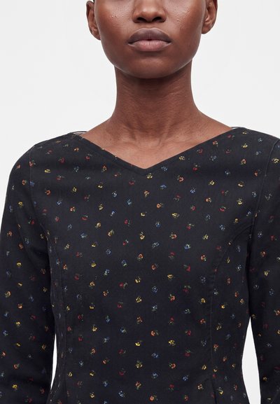 Personne portant un haut noir à manches longues avec un motif tacheté coloré subtil et un col en V large.