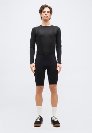 Mladý muž stojící před světlým pozadím, oblečený v černém dlouhém rukávu, černých cyklistických kraťasech s laclem, bílých ponožkách a černých teniskách.