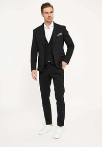 Schwarzer Dreiteiler mit einem weißen Hemd, ergänzt durch ein Einstecktuch, kombiniert mit weißen Sneakers. Schlanker Schnitt und maßgeschneiderte Gestaltung.