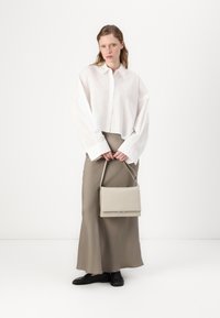Weißes oversize Hemd mit langen Ärmeln, kombiniert mit einem beigen Maxikleid. Eine kleine beigen Handtasche wird vorne gehalten. Schwarze Schuhe vervollständigen das Outfit.