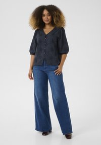 Blusa blu navy con scollatura a V e texture floreale, abbinata a jeans a gamba larga blu. La blusa presenta maniche corte a sbuffo e bottoni.