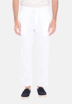 Uomo che indossa pantaloni bianchi con coulisse e scarpe slip-on nere, in piedi davanti a uno sfondo bianco semplice.