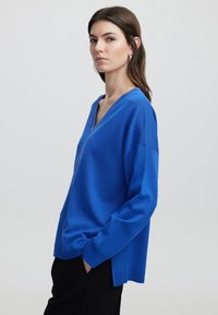 Blaues Strickpullover mit V-Ausschnitt, langen Ärmeln, geripptem Saum und seitlichen Schlitzen, getragen mit schwarzen Hosen. Weiche Textur und lockere Passform.