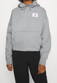 Felpa grigia con cappuccio regolabile, tasca a marsupio e una patch rossa con il logo che presenta il profilo di una figura che salta. Materiale in cotone morbido.