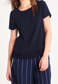 Navy short-sleeve top with a round neckline, featuring a pleated fabric accent at the hem. Paired with navy striped wide-leg pants.