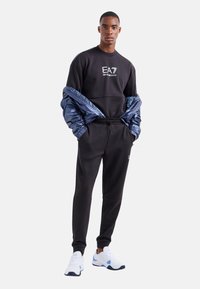 Sudadera negra con logo, combinada con pantalones de chándal negros. Una chaqueta azul brillante caída sobre los hombros y zapatillas blancas con acentos azules.