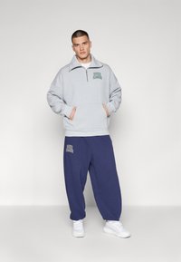 Sudadera gris con cremallera y un logo verde, combinada con pantalones de chándal holgados azul marino que tienen un logo, y terminada con zapatillas blancas.