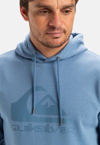 Ljusblå hoodie med en stor grafisk logga i en mörkare nyans. Har en dragsko i luvan och en slät tygtextur.