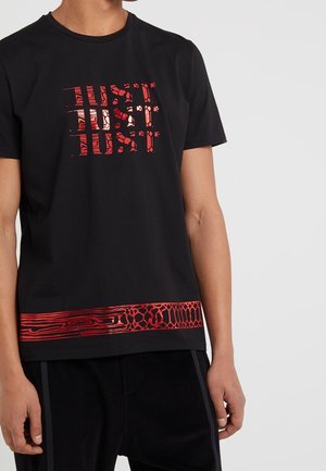 Schwarzes kurzärmeliges T-Shirt mit rotem, strukturiertem Schriftzug "JUST", der dreimal auf der Brust wiederholt wird, und rotem gemustertem Band oberhalb des Saums, getragen von einer Person.