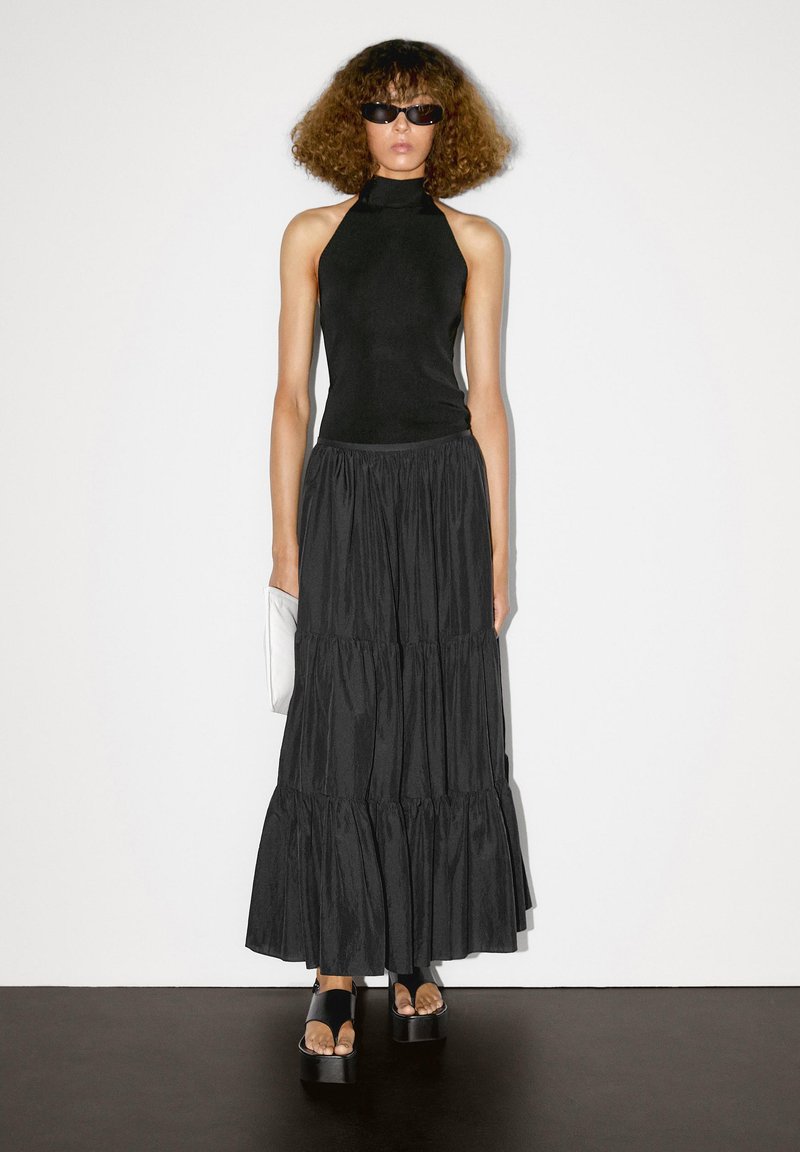 Top a fascia nera con un corpetto aderente, abbinato a una gonna maxi nera a più strati in tessuto leggero. Sandali con zeppa e una clutch bianca completano il look.