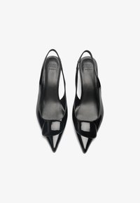 Fekete lakk bőr slingback magassarkú cipők hegyes orral és dekoratív pánttal az elején. Simább textúrájú és minimalista formatervezésű.