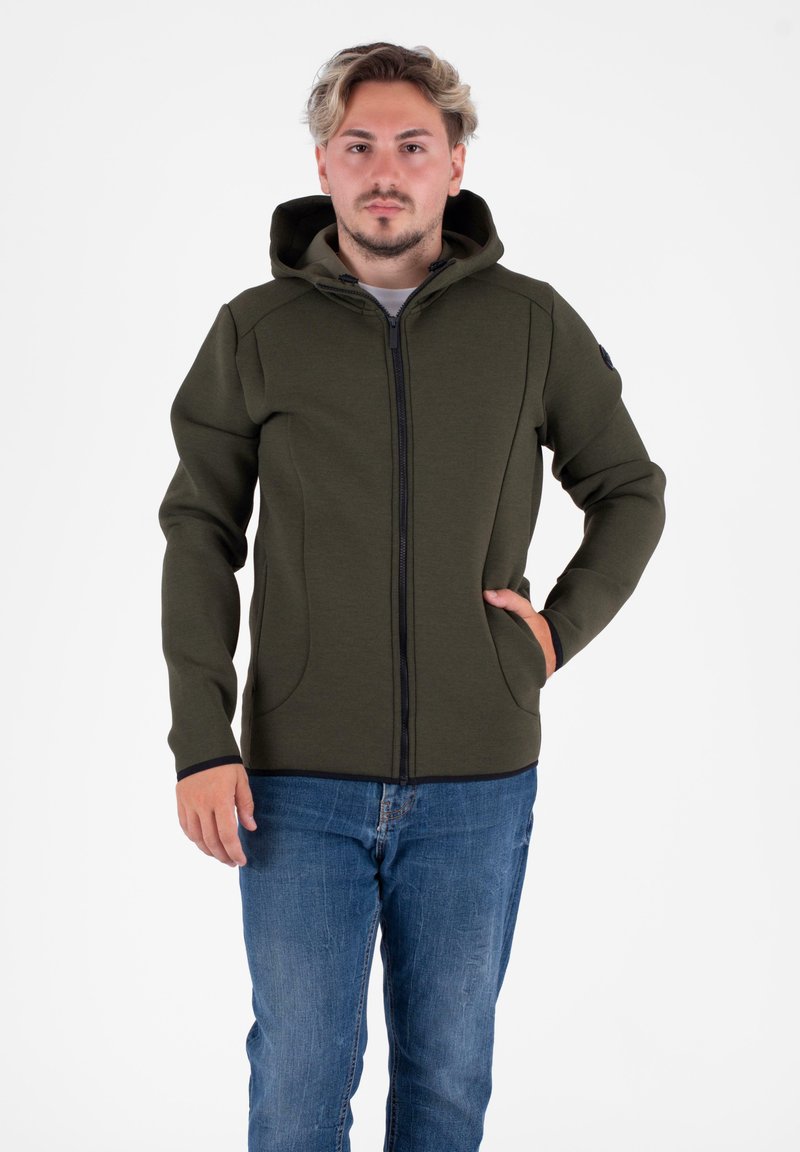 Felpa con zip di colore verde oliva, dotata di cappuccio, maniche raglan, tasche laterali e dettagli di cucitura a contrasto, abbinata a jeans blu.