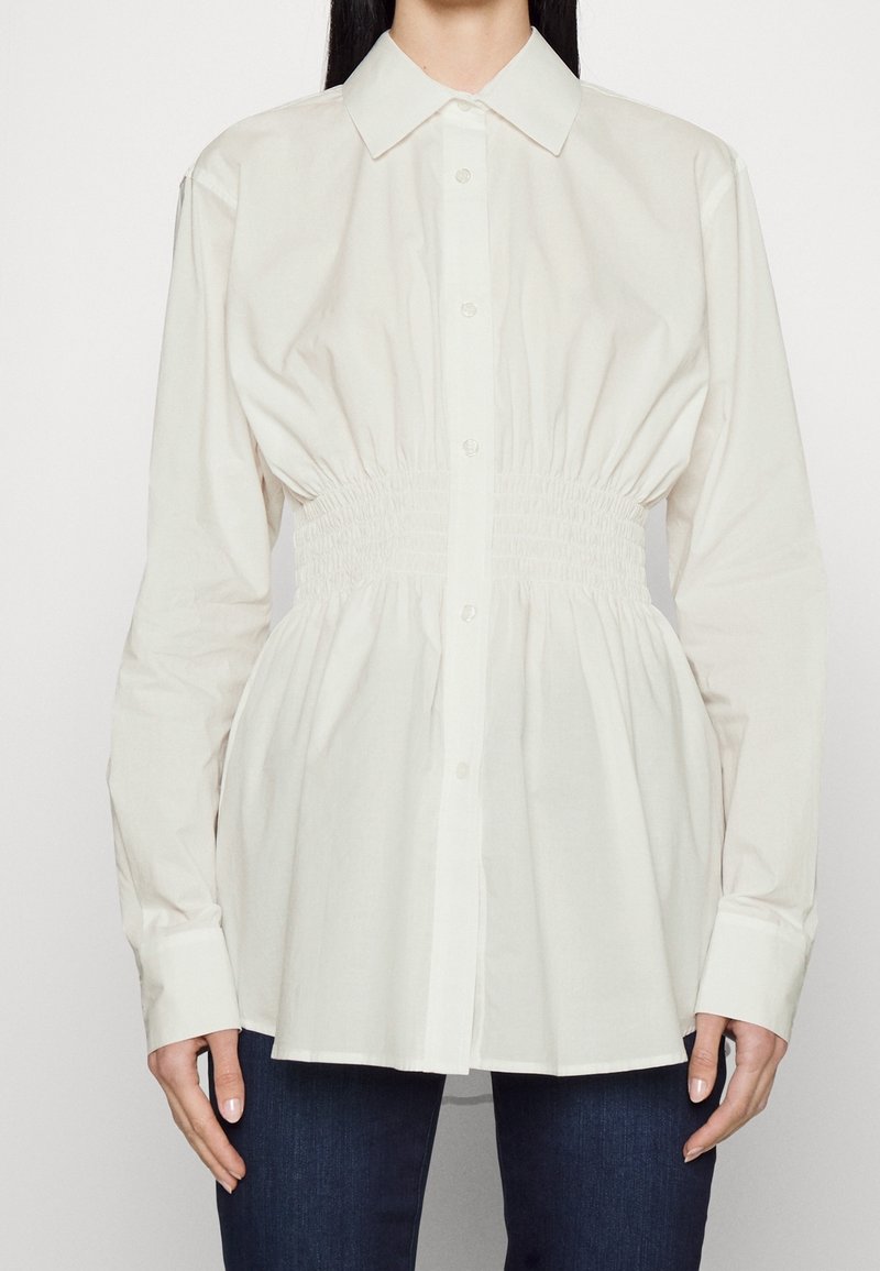 Femme portant une chemise blanche à manches longues boutonnée avec une taille élastiquée froncée et un pantalon bleu foncé.