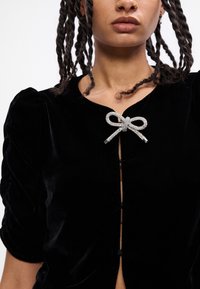 Blusa in velluto nero con maniche a sbuffo, caratterizzata da un'evidente chiusura a fiocco in strass argentati al collo e un dettaglio dei bottoni sul davanti.