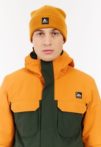 Svetlooranžová a tmavozelená nepremokavá bunda s dvomi náprsními vreckami a nastaviteľnou kapucňou; v kombinácii s horčicovožltou pletenou čiapkou.