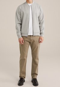 Šedá bomber bunda na zip přes bílý tričko, doplněná khaki kalhotami a černými botami, kterou nosí osoba stojící na neutrálním pozadí.