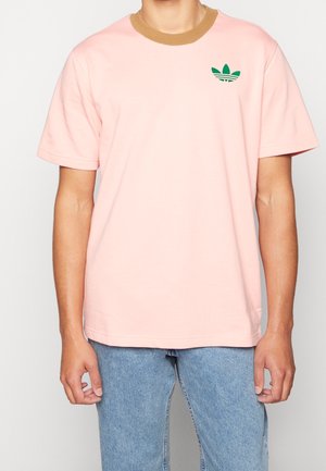 Camiseta de algodón de color rosa claro con un cuello acanalado marrón y un logo de trébol verde de Adidas en el pecho. Mangas cortas y un ajuste holgado.