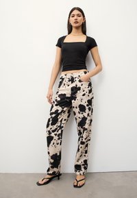 Schwarzes Kurzarmoberteil kombiniert mit beigen Hosen mit schwarzem Spritzmuster. Flache Sandalen vervollständigen das Outfit.