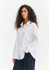 Biała koszula zapinana na guziki z kołnierzykiem, z czerwonym akcentem na lewym ramieniu. Długie rękawy i luźny krój. Połączona z ciemnym dżinsem.