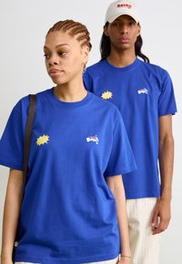 Twee personen dragen levendige blauwe katoenen t-shirts met ronde hals en korte mouwen. Opvallende graphics omvatten logo's en kleurrijke accenten.