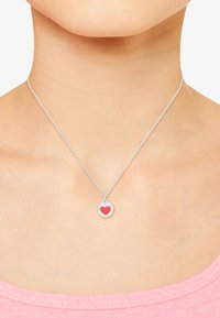 Collar de plata con un colgante redondo que presenta un diseño de corazón rojo, llevado en un cuello desnudo, acompañado de una camisa rosa.