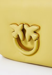 Borsa a tracolla gialla in pelle con un accento in metallo dorato a forma circolare con dettagli a forma di foglia, che mostra una texture liscia.