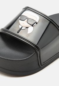 Sandale negre tip slide cu talpă texturată și o curea lucioasă cu o față de desen animat în relief care poartă ochelari de soare și cravată.