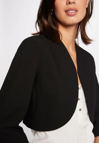 Schwarze cropped Jacke mit rundem Halsauschnitt und ohne Verschlüsse. Weicher Stoff mit minimalistischem Design und ohne sichtbare Muster.