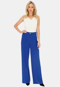 Biała bezrękawnikowa bluzka zestawiona z niebieskimi spodniami o wysokim stanie i szerokich nogawkach, z złotym guzikiem, gładkim materiałem i luźnym krojem.