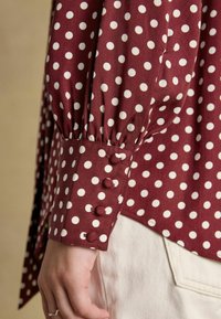 Burgundy polka dot shirt with white dots, featuring gathered sleeve, buttoned cuff with three matching fabric-covered buttons.