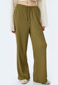 Pantaloni larghi in verde oliva, con texture e vita regolabile. Il tessuto sembra leggero e traspirante, con sottili linee verticali.