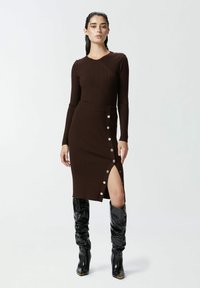 Abito in maglia a coste marrone con maniche lunghe, dettagli con bottoni laterali e uno spacco alto fino alla coscia. Abbinato a stivali neri al ginocchio con punte dorate.