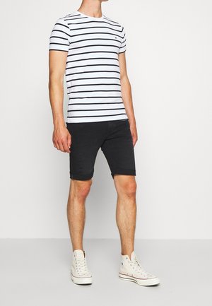 T-shirt bianca a maniche corte con strisce orizzontali nere, abbinata a pantaloni shorts in denim neri con dettagli strappati e orli arrotolati. Sneakers bianche.