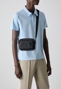 Svart zip-försett crossbody-väska med präglad logotyp, bärs över en ljusblå skjorta med krage, i kombination med beige byxor. Texturerad tygdetalj synlig.