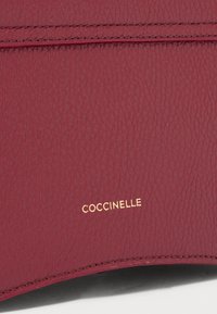 Portofel din piele texturată maronie cu branduirea aurie "Coccinelle"; prezintă cusături fine și o suprafață netedă.