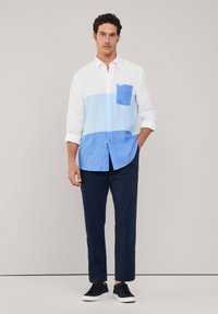 Camicia button-up a righe azzurro chiaro e bianco con taschino sul petto, abbinata a pantaloni navy e sneakers nere. Tessuto di cotone, design informale.