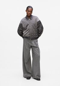 Bomber grigio con motivi sottili, chiusura a zip e polsini a coste; abbinato a pantaloni larghi grigi e scarpe nere.