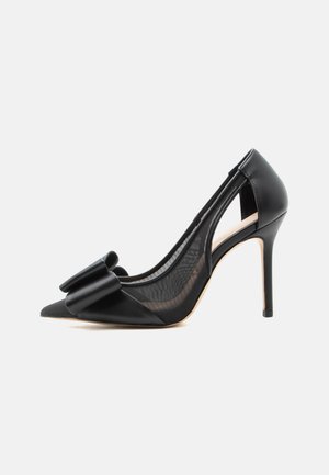 Chaussure noire à talons hauts avec bout pointu, panneaux latéraux transparents et grand nœud décoratif à l'avant, vue de profil droit.