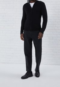 Svart zip-up tröja med ribbade ärmslut och nederkant, tillverkad med svarta byxor och svarta skor, som står mot en vit tegelbakgrund.