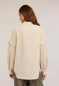 Beige bluse med lange puffærmer, udstyret med bundet manchetter og en klassisk krave. Fremstillet af glat stof, med en let forlænget bagkant.