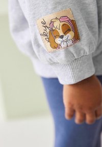 Szara bluza z miękkiej tkaniny, z naszywką w kształcie kreskówkowego psa w beżowym kolorze z różowymi akcentami na rękawie. Długie rękawy z ściągaczami.