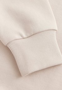 Tejido beige con un puño acanalado, que muestra una textura suave y un sutil patrón de punto. Enfócate en el ajuste a medida de la manga y la consistencia del color.