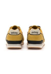 Un par de zapatillas amarillas y negras con medias suelas blancas, vistas desde la parte trasera, mostrando el logo "mtng" en las pestañas del talón.