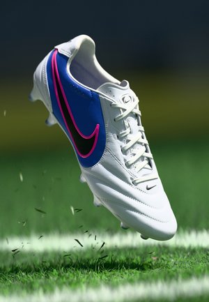 Chaussure de soccer blanche avec des accents bleus et roses, comportant un swoosh noir distinctif, des lacets, et une surface texturée, flottant au-dessus de l'herbe.