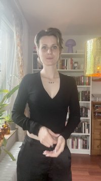 Black long-sleeve top with a deep V-neck, featuring a ribbed texture. The individual is wearing black pants, with a light background and bookshelves.