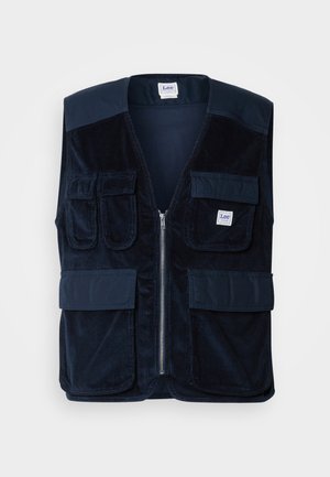 Colete de corduroy azul-marinho com fecho zip, quatro bolsos frontais e um rótulo. Apresenta um design de ombro liso e um acabamento texturizado.