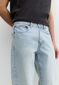Helles blaue Denim-Jeans mit lockerer Passform, sichtbaren Nähten, fünf Taschen und silbernem Zubehör. Getragen mit einem dunkelgrauen kurzärmeligen Shirt.