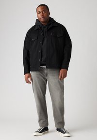 Chaqueta de mezclilla negra sobre una sudadera gris, combinada con jeans grises y zapatillas negras. La chaqueta cuenta con cierres de botón y bolsillos frontales.