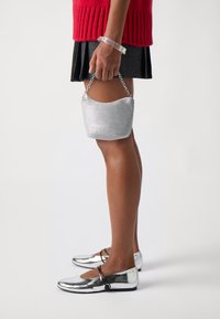 Stříbrná kovová kabelka s zakřiveným tvarem a řetízkovým uchem, doplněná lesklými balerínami a texturovanou černou sukní. Viditelný červený pletený top.