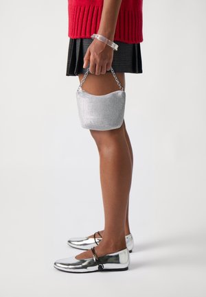 Zilverkleurige metalen handtas met een gebogen vorm en kettinghengsel, gecombineerd met glanzende platte schoenen en een gestructureerde zwarte rok. Rode gebreide top zichtbaar.